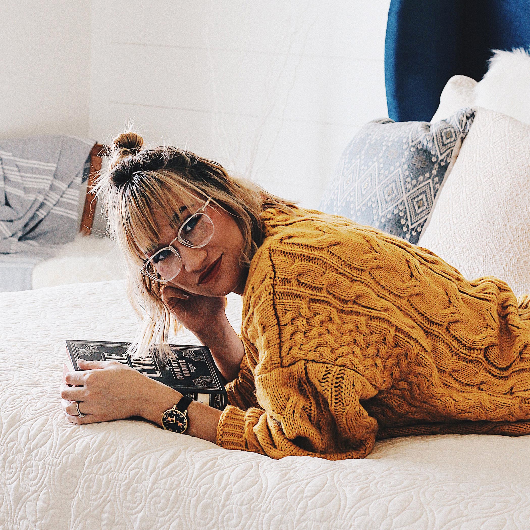 Woman in a mustard cable-knit sweater wearing glasses, holding a book while lying on a bed.