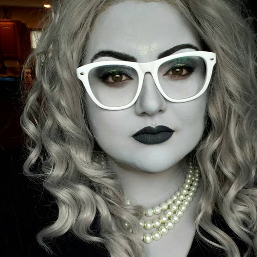 Woman with silver hair and white glasses wearing a pearl necklace, dark makeup, and gray face paint.