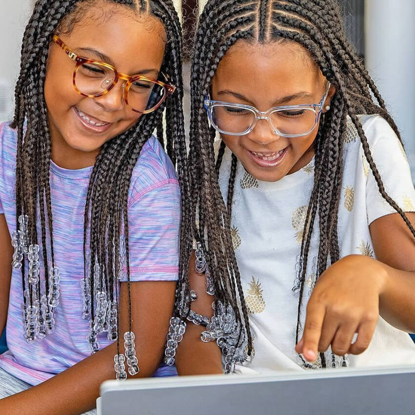 Two children with glasses wearing colorful shirts, smiling and looking at a tablet together.