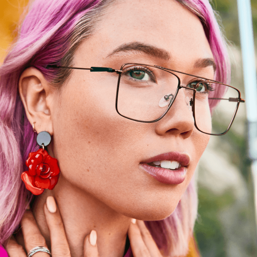 Woman with purple hair wearing rectangular wire glasses and red rose earrings.