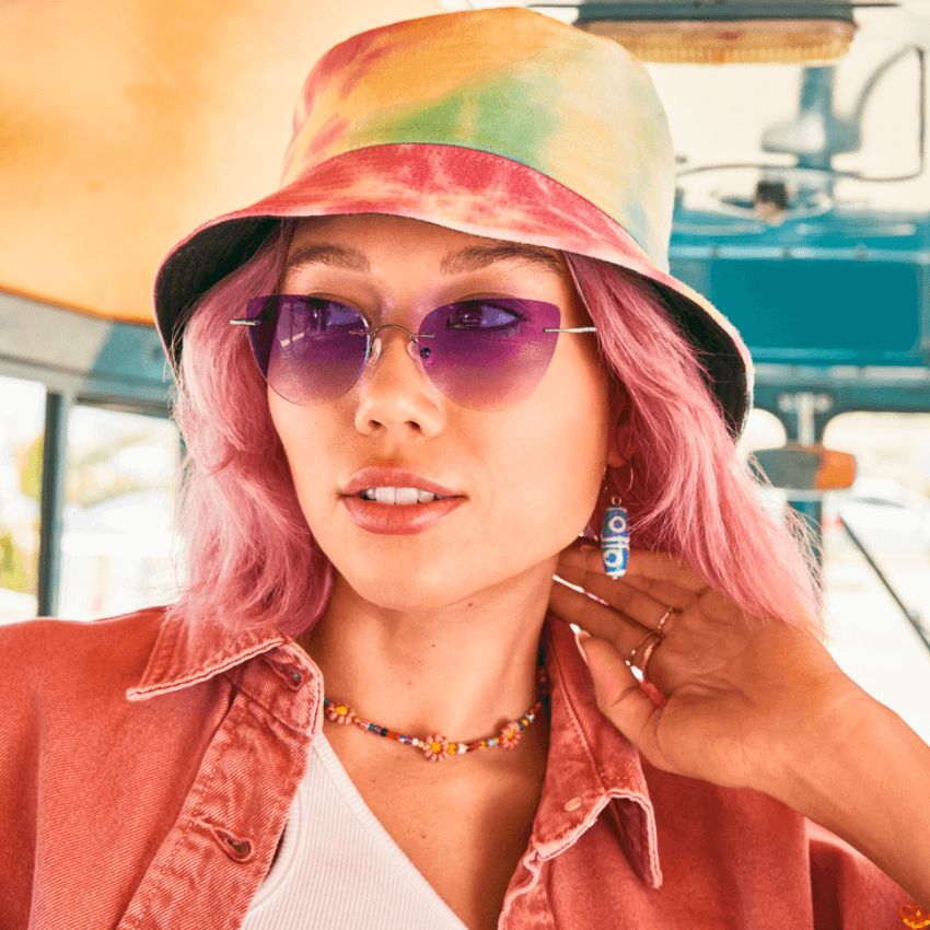 Woman wearing purple-tinted sunglasses, a tie-dye bucket hat, and a pink shirt with a floral necklace.