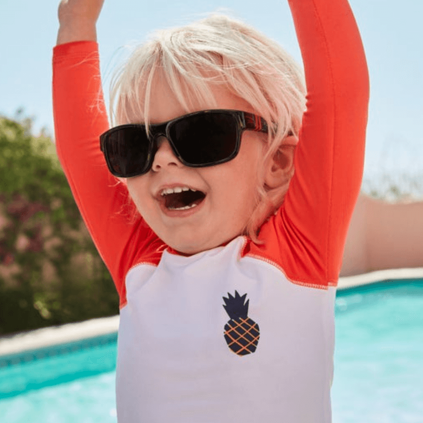 Child wearing black sunglasses and a white rash guard with orange sleeves and a pineapple design.
