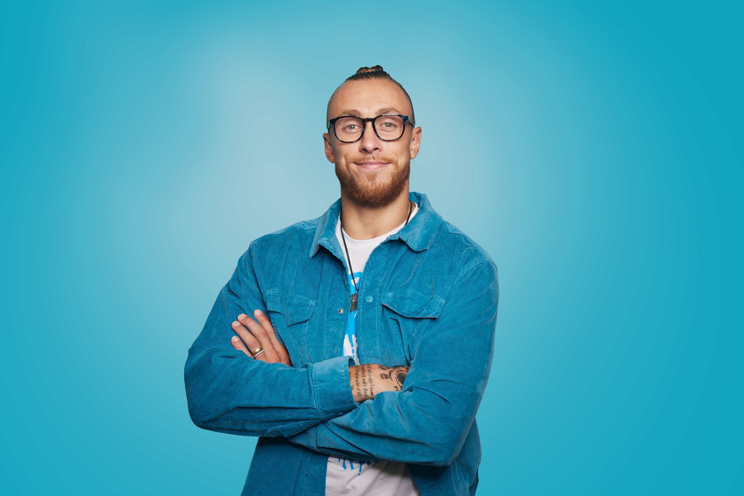 Person wearing glasses and blue denim jacket with arms crossed against a plain blue background.