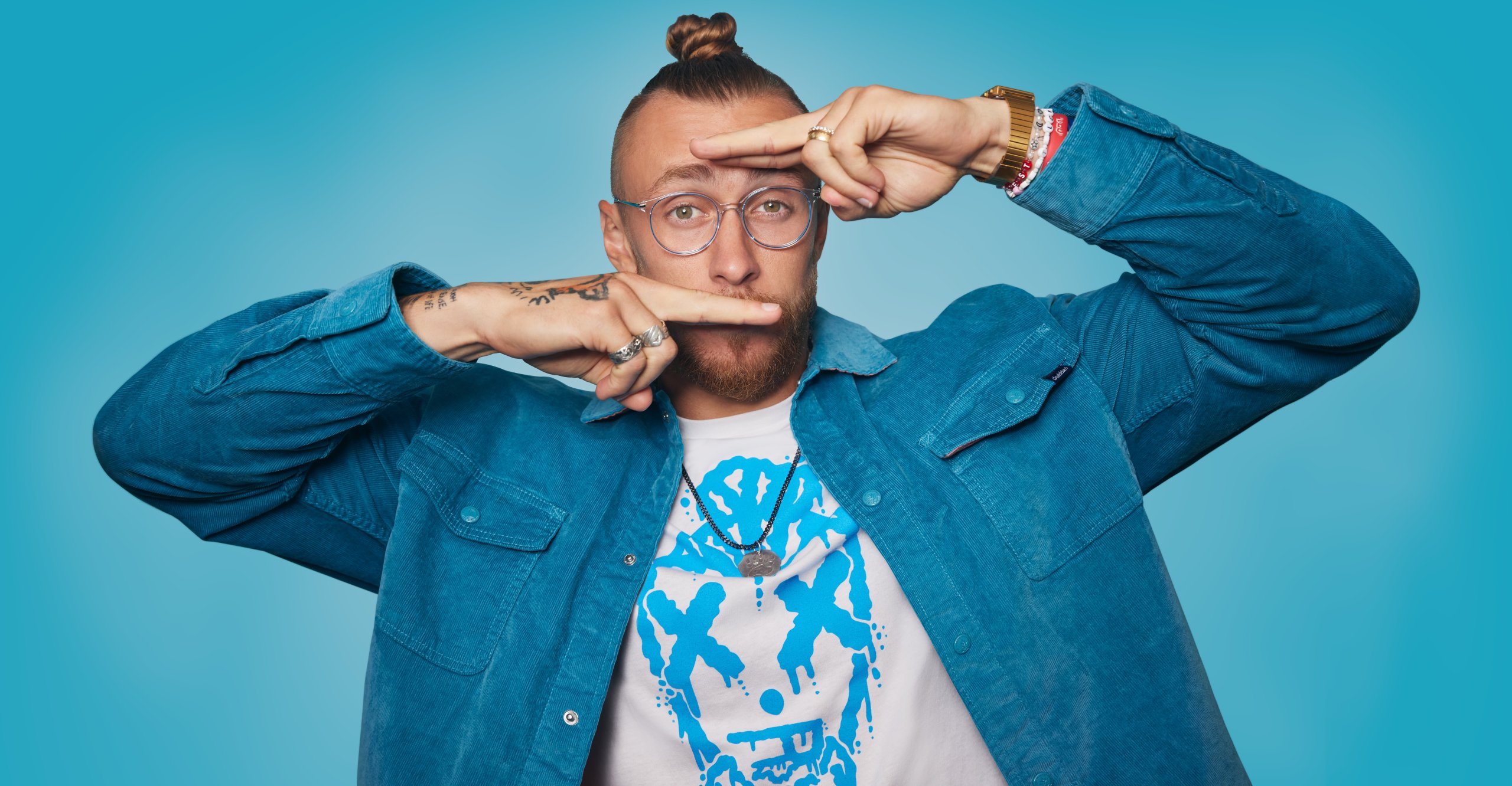 Man wearing blue jacket and glasses, white t-shirt with blue graphic, making a framing gesture with hands.