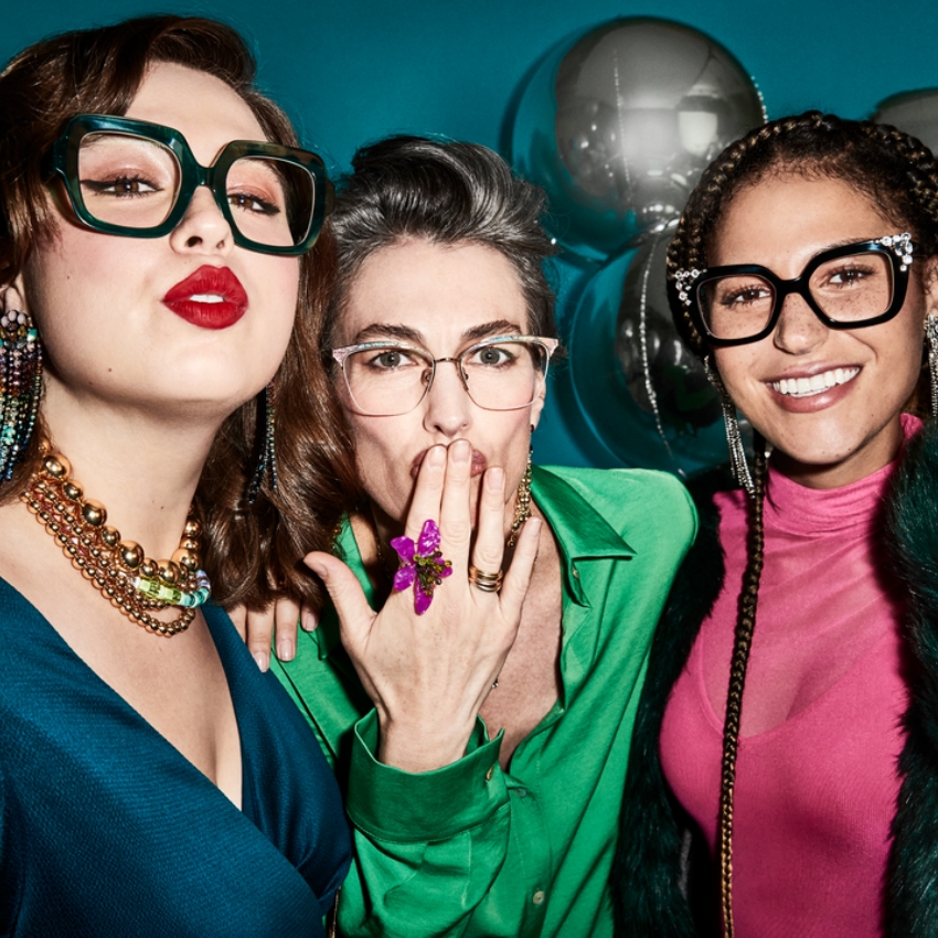 Three women with glasses, colorful earrings, and bold outfits. Central person wears a ring with a purple flower.