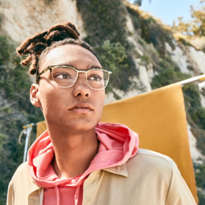 Person wearing stylish, clear-framed glasses and a pink hoodie under a beige jacket.