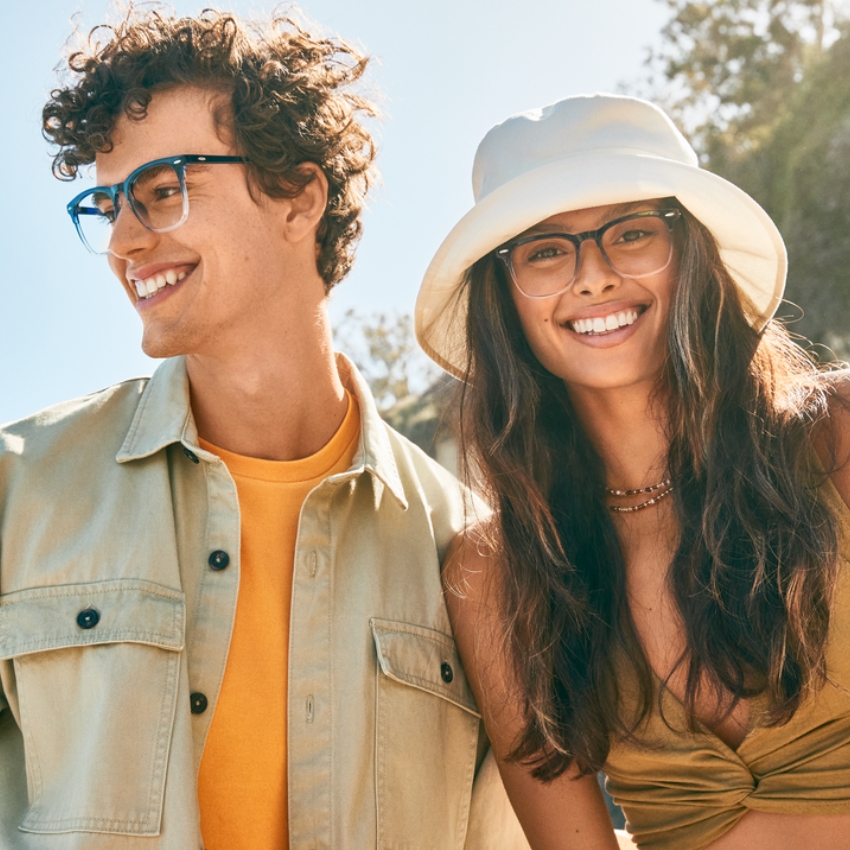 Man and woman smiling, both wearing stylish eyeglasses; woman also wearing a white bucket hat.