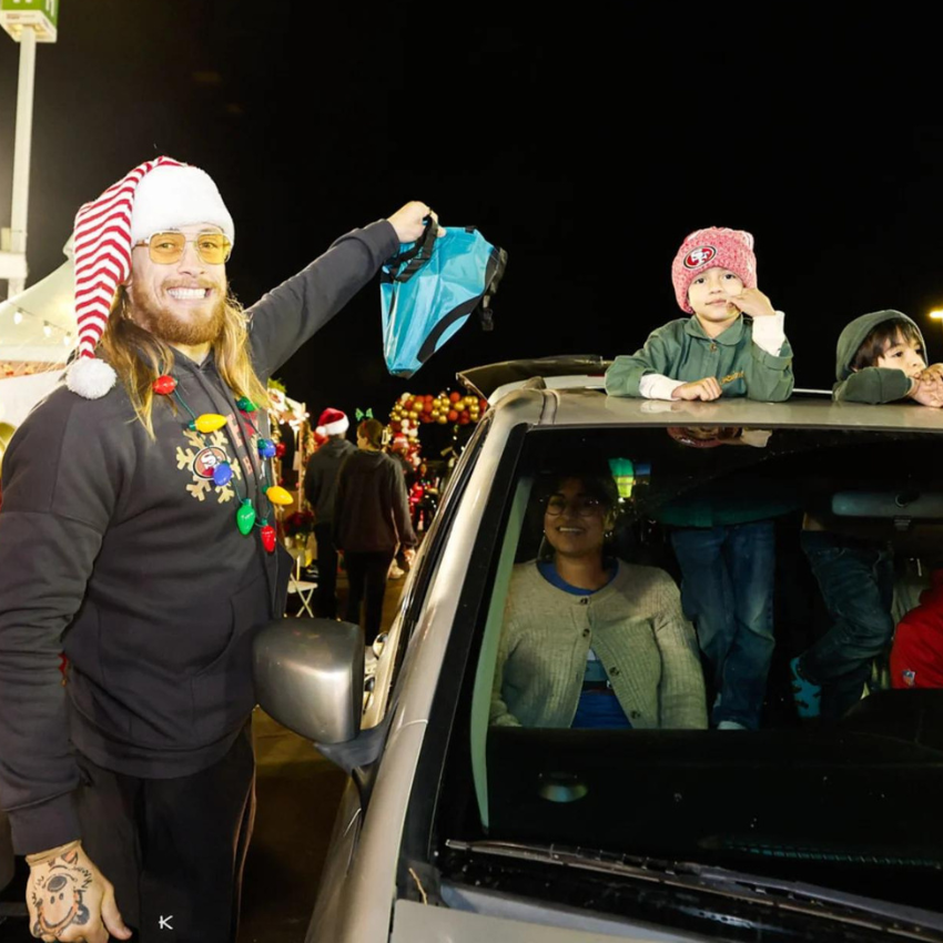 Man in Santa hat with Christmas lights on hoodie giving a gift bag to children in car's sunroof.