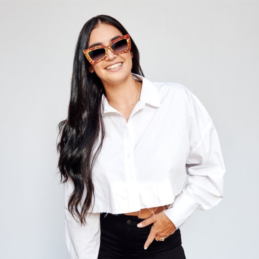 Woman wearing tortoiseshell patterned square sunglasses, white cropped blouse, and black pants, smiling.