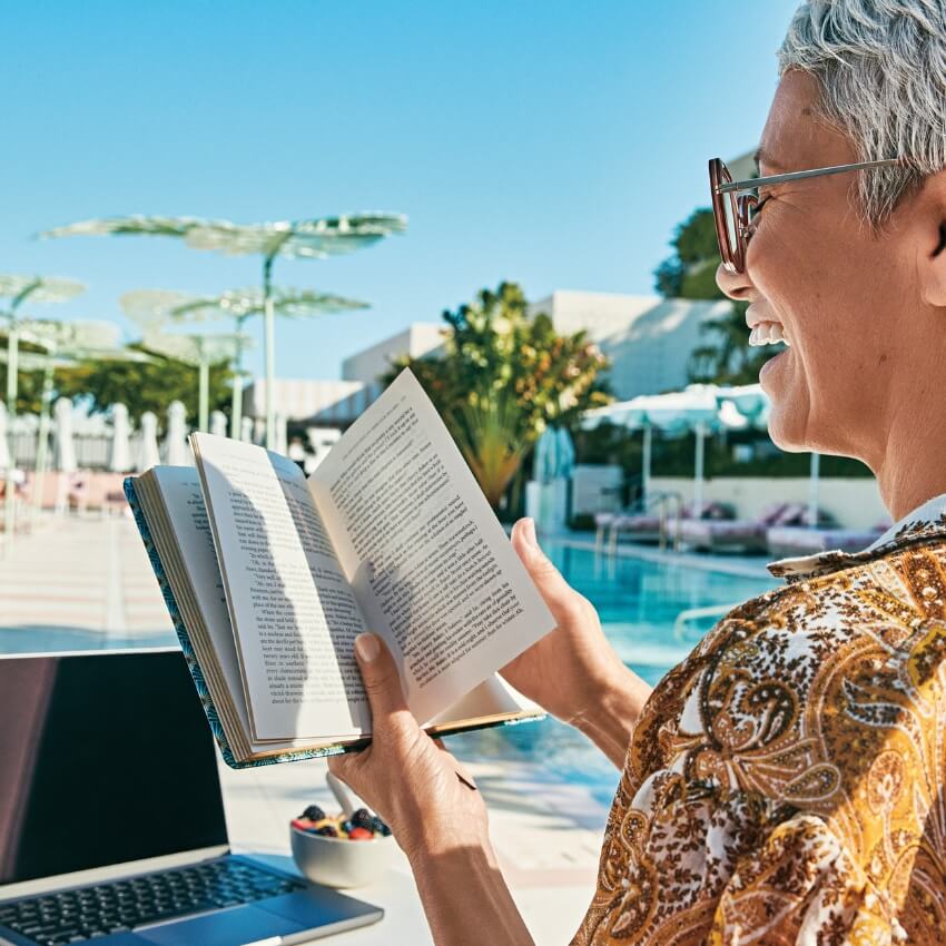 Person in patterned shirt reading a book near a pool, with a laptop and a bowl of fruits on the table.