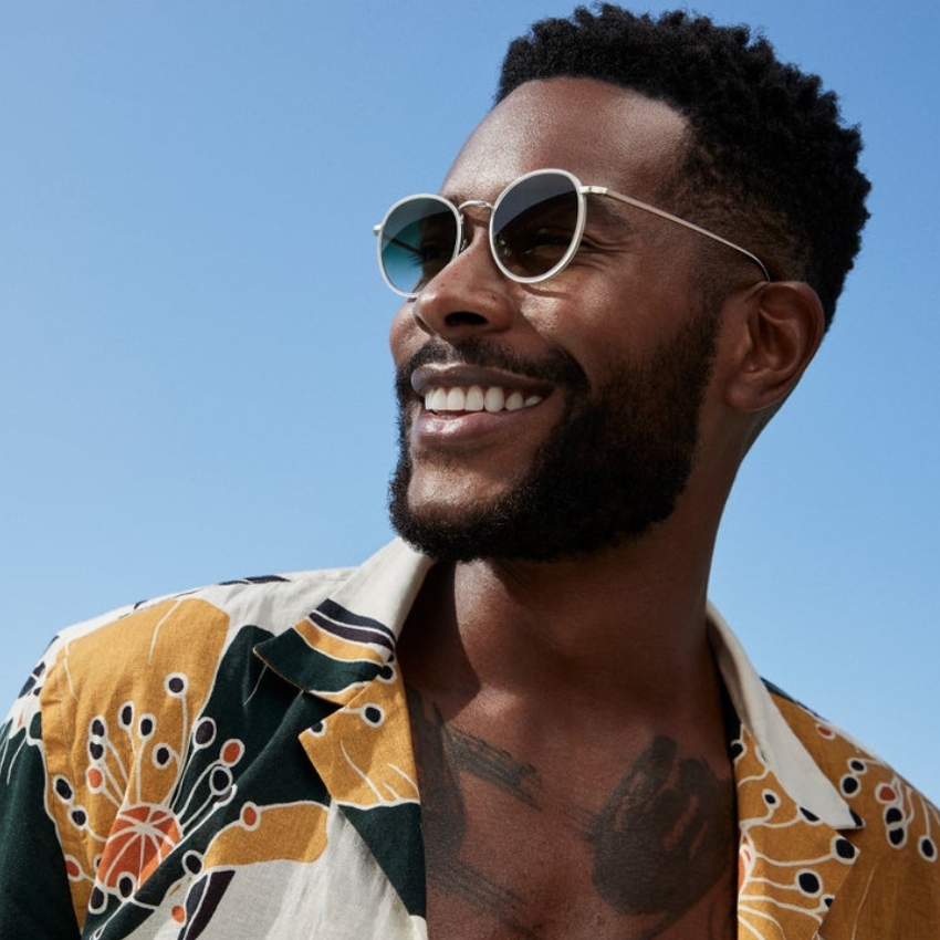 Man wearing round silver sunglasses and a yellow, green, and white floral shirt with black trim.