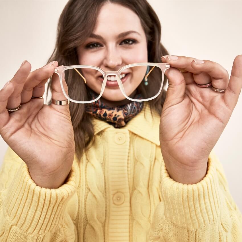 Woman holding clear eyeglass frames in front of her face, wearing a yellow sweater and patterned scarf.