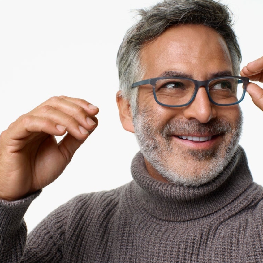 Man adjusting dark-rimmed glasses, wearing a gray turtleneck sweater, and smiling.