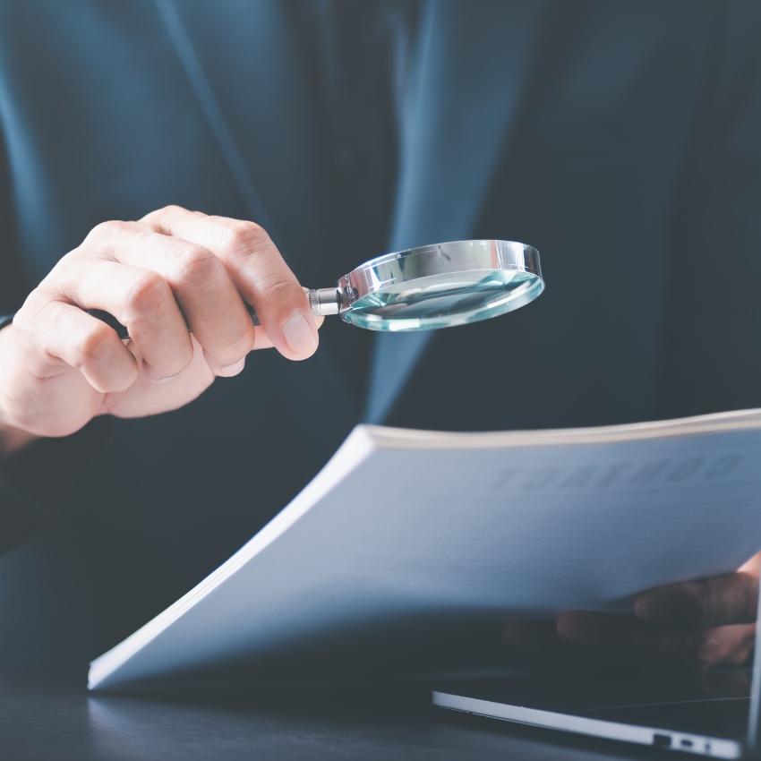 Hand holding a magnifying glass over a document. The document text appears to be "CONTRACT."