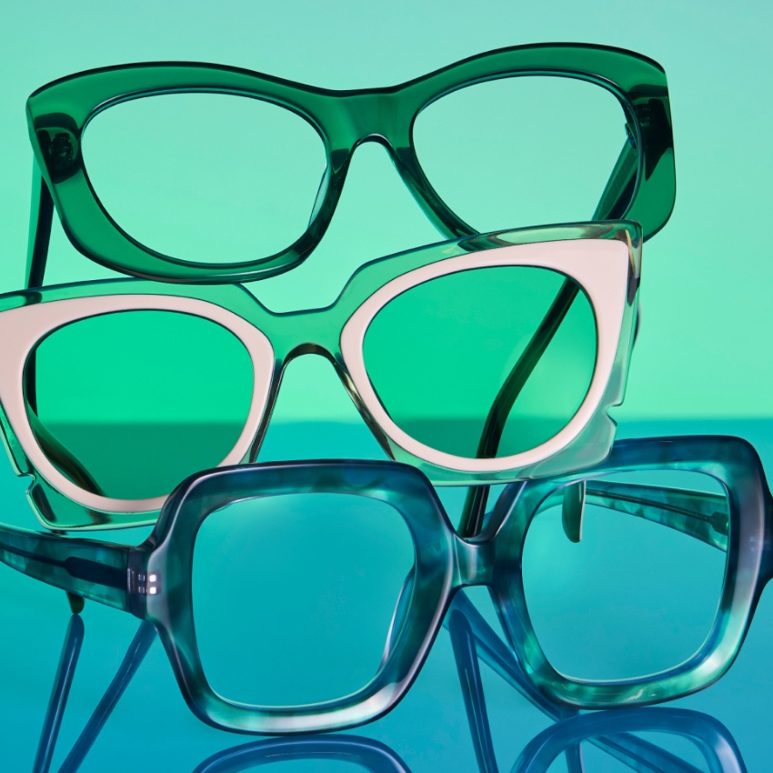 Three pairs of bold eyeglasses with green, white, and blue frames stacked on a reflective surface.