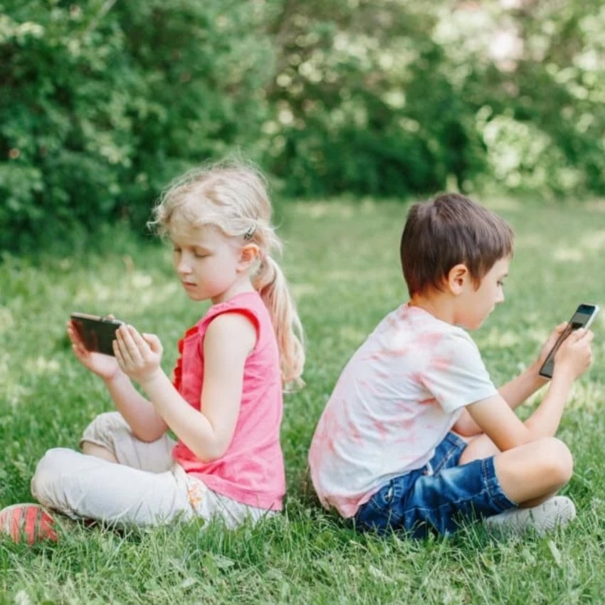Two kids sit on grass back-to-back, each using a smartphone.