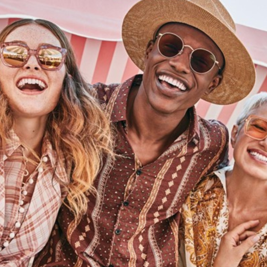 Group of friends wearing stylish sunglasses, patterned shirts, and a straw hat, smiling and laughing.