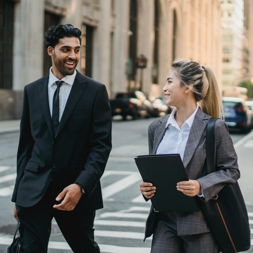 Two professionals walking and talking, both dressed in suits. The woman is holding a black folder.