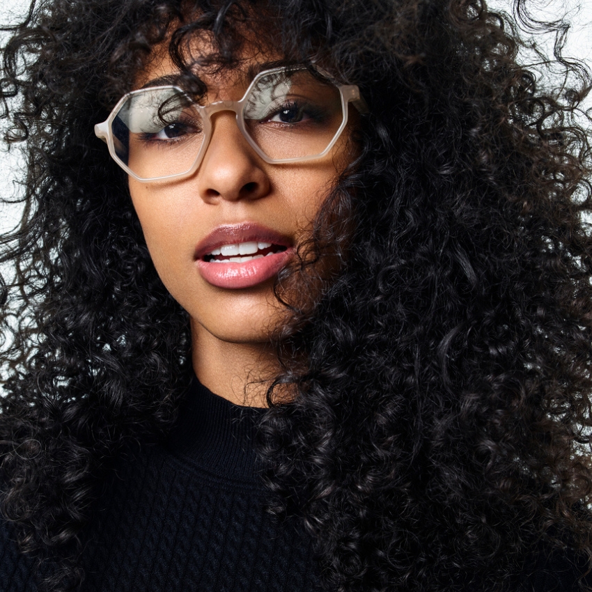 Curly-haired model wearing geometric, translucent eyeglasses and a black textured top.