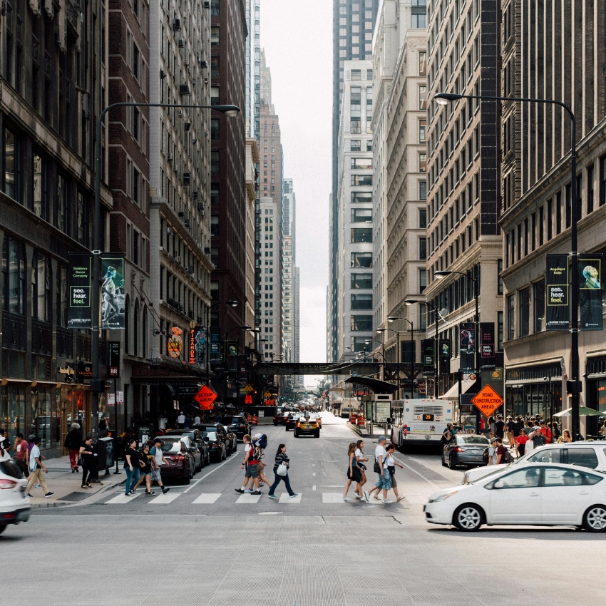 Busy urban street with people crossing, tall buildings, cars, and "Road Construction Ahead" signs visible.