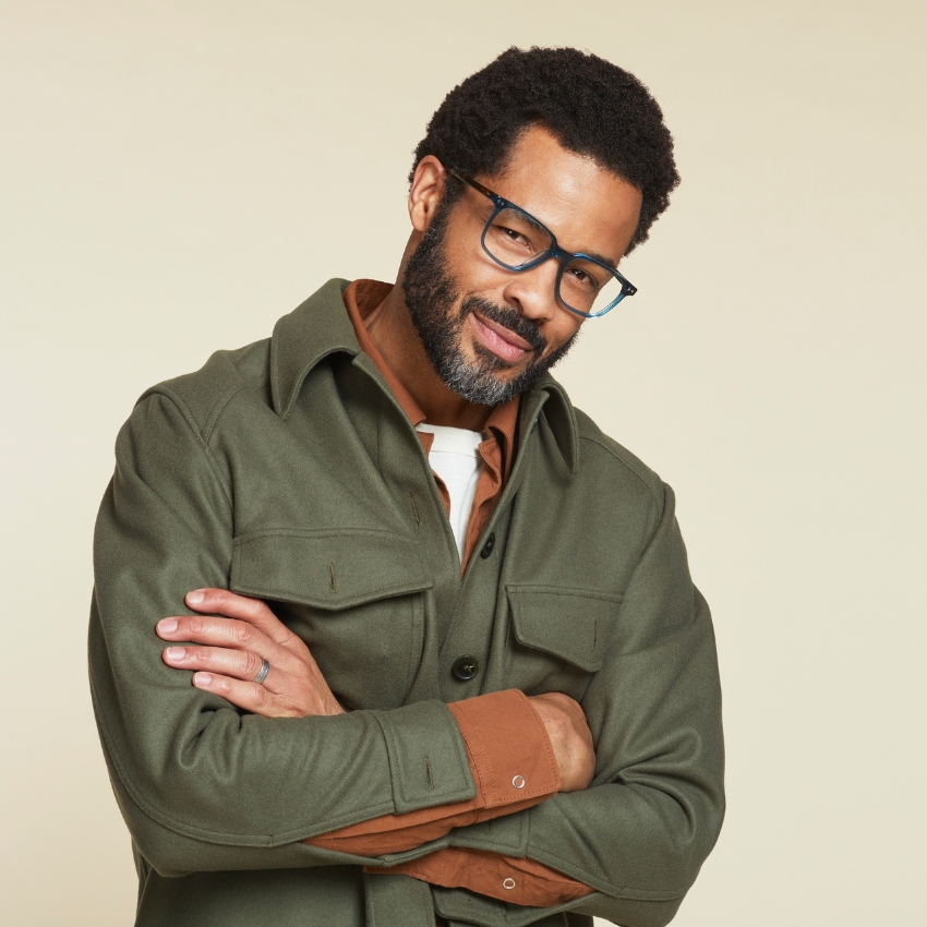 Man wearing an olive green jacket with a brown collar, blue-framed glasses, and a white undershirt.