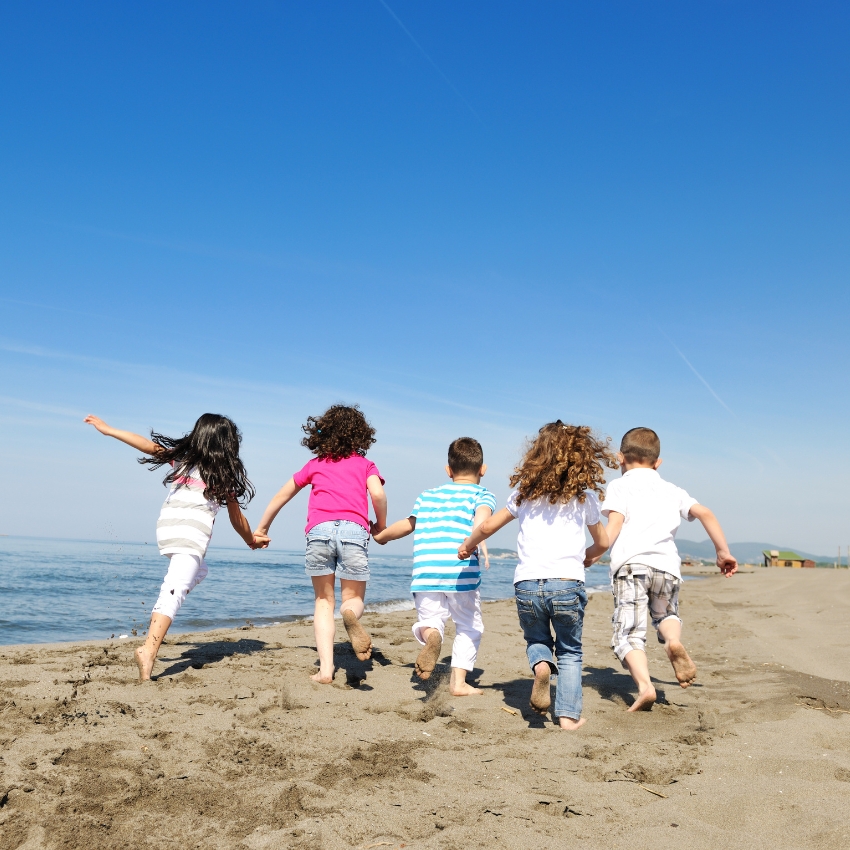 Children holding hands running on a beach toward the ocean under a clear blue sky.